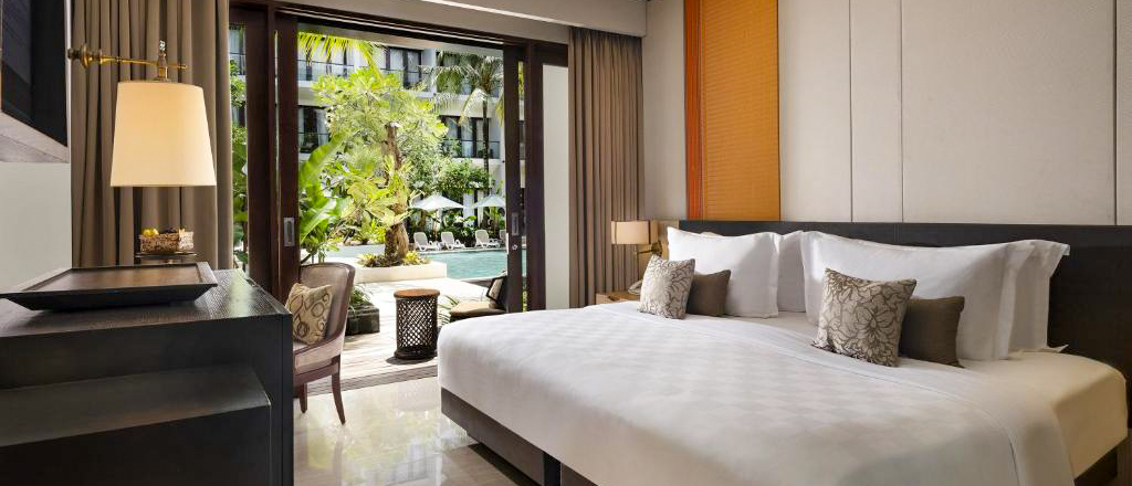 Luxurious hotel room in Kuta with a large bed and open doors to a tropical swimming pool.