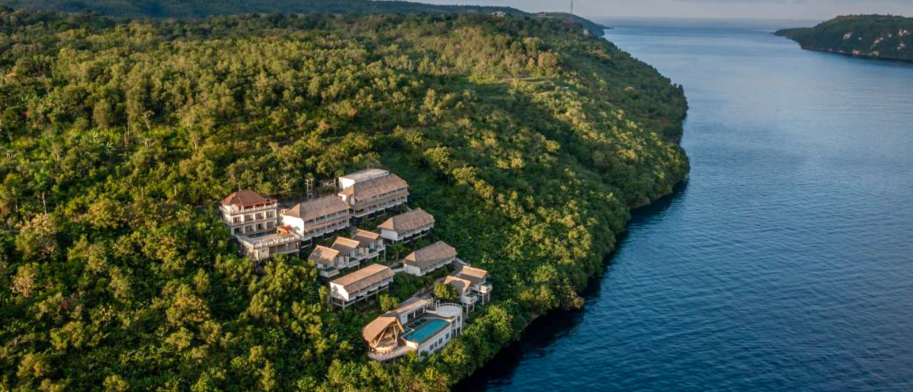 Luxury resort buildings and pools on a lush cliff overlooking the calm blue ocean, Nusa Penida.