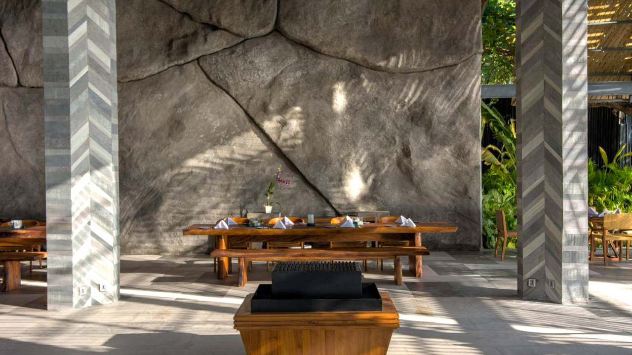 A serene outdoor dining area at a Lombok resort, featuring a long wooden table and textured wall.