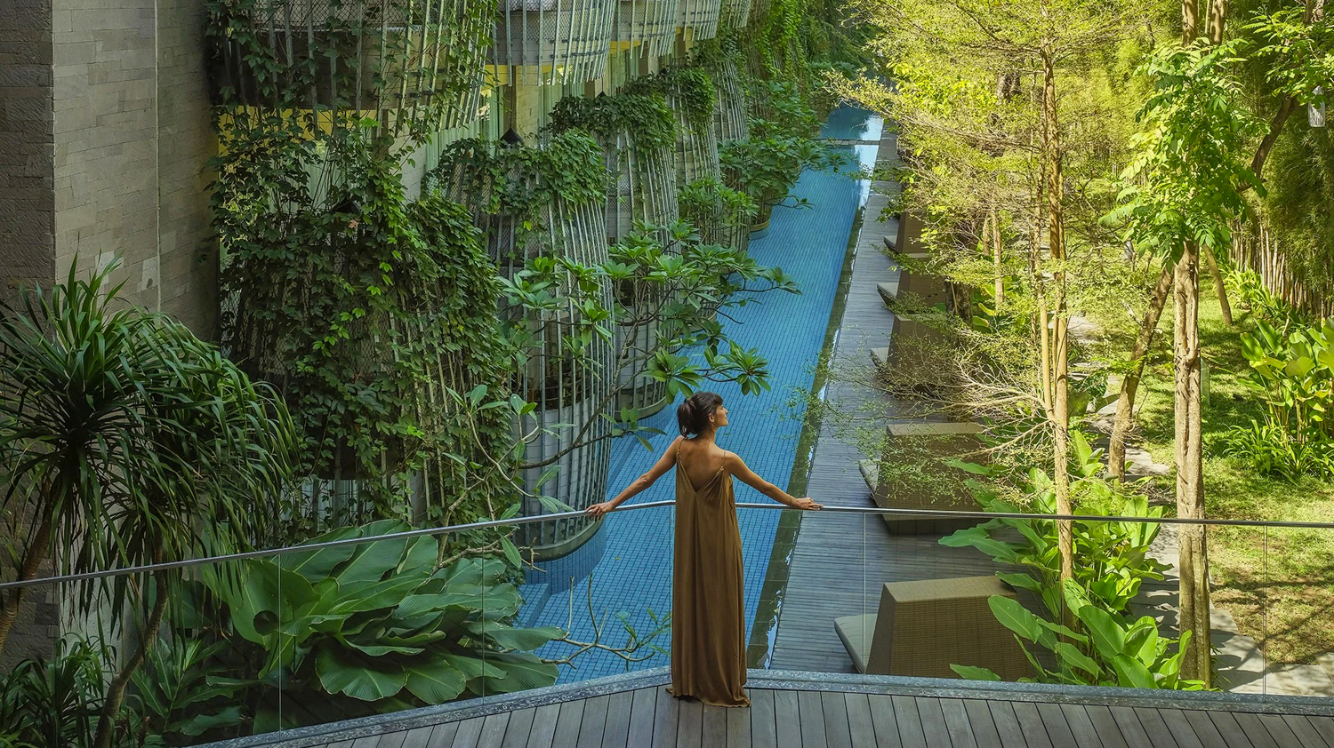 Woman in a long dress on a wooden deck overlooks a serene resort pool and lush greenery.