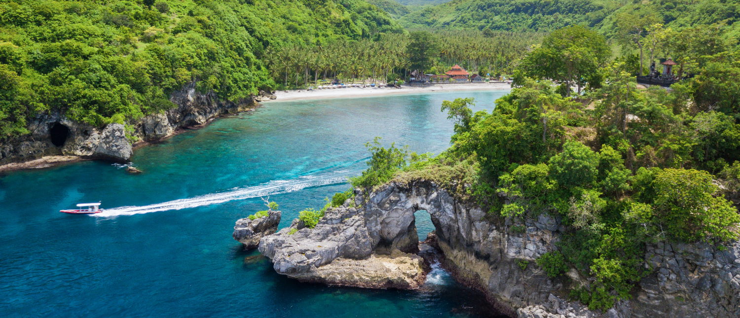 Crystal Bay, Nusa Penida, with a boat speeding past a natural rock arch in clear waters.