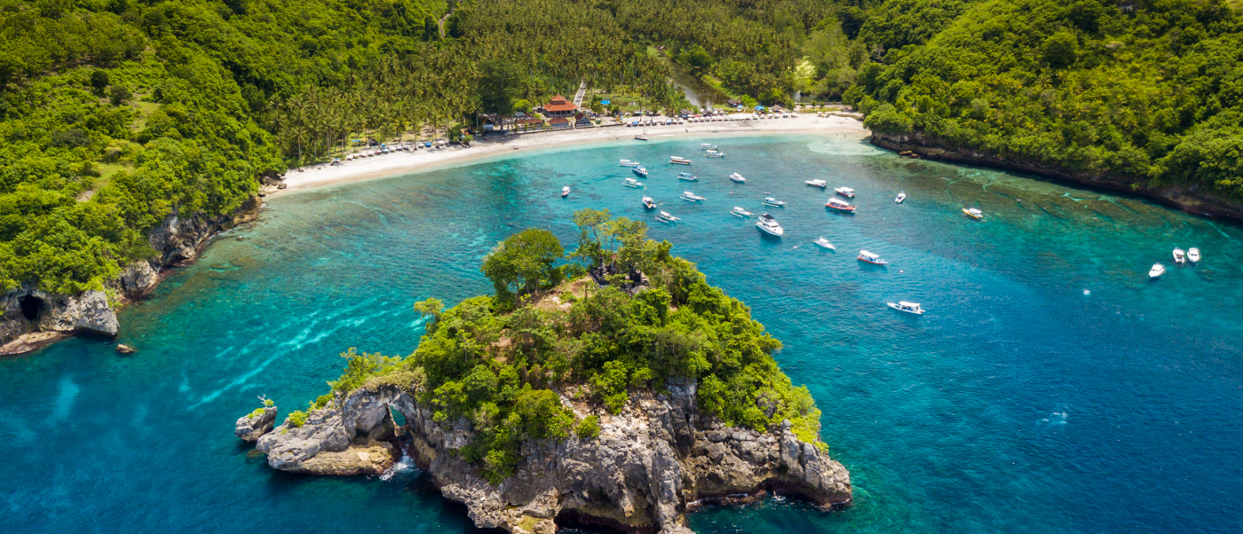 Aerial view of Crystal Bay, Nusa Penida, showing a rocky island, clear turquoise water, and many boats.