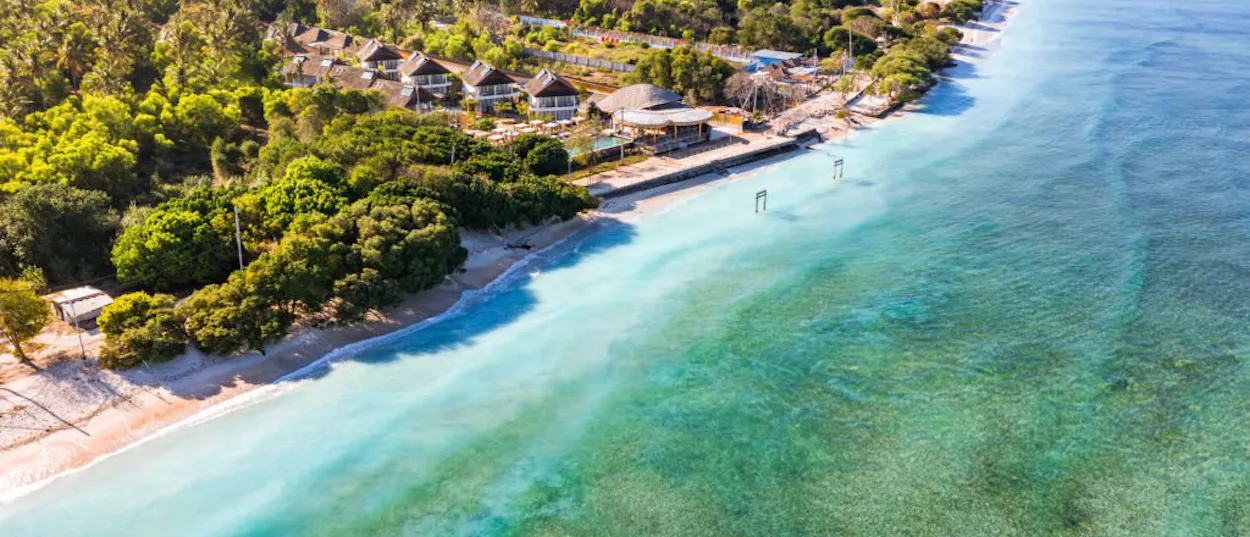 Gili Trawangan's serene coastline featuring clear turquoise ocean, a sandy beach, and tropical bungalows nestled in trees.