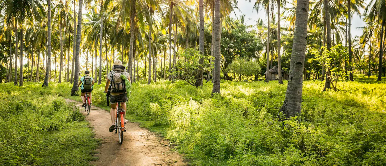 Two people cycle on a dirt path through a tropical palm forest on Gili Trawangan island.