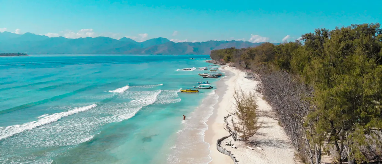 Aerial view of Gili Trawangan beach with turquoise water, boats, and lush trees under a clear sky.