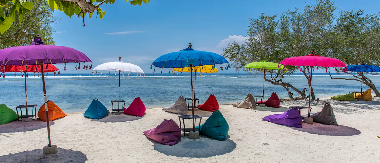 Colorful bean bags and parasols line a vibrant sandy beach with clear blue water on Gili Trawangan.