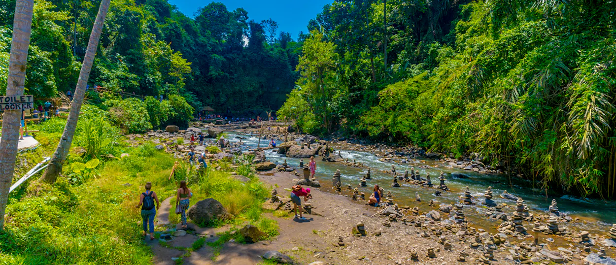 Visitors enjoy the scenic river and stacked stones amidst dense tropical jungle in Ubud, Bali.
