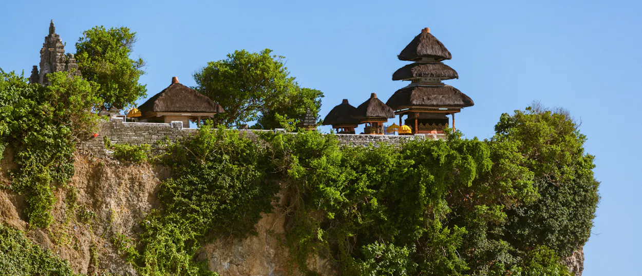 Traditional Balinese temple buildings with thatched roofs sit atop a dramatic, green-covered cliff at Uluwatu.