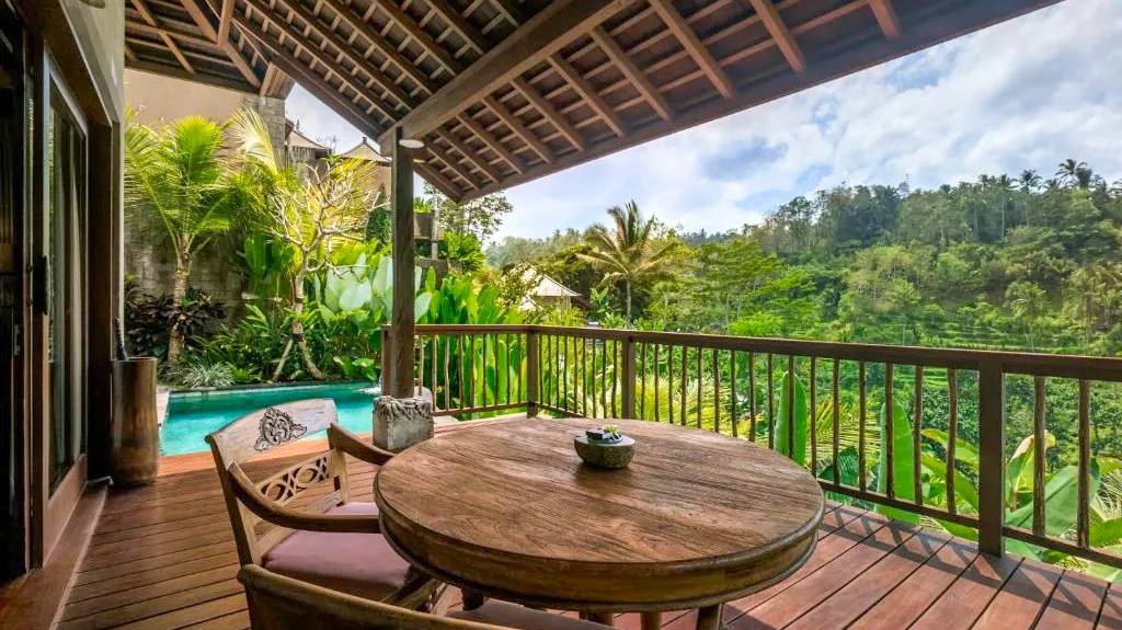 A private wooden balcony with a table and chairs, overlooking a lush Bali jungle and pool.
