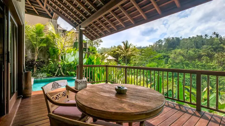 A private wooden balcony with a table and chairs, overlooking a lush Bali jungle and pool.