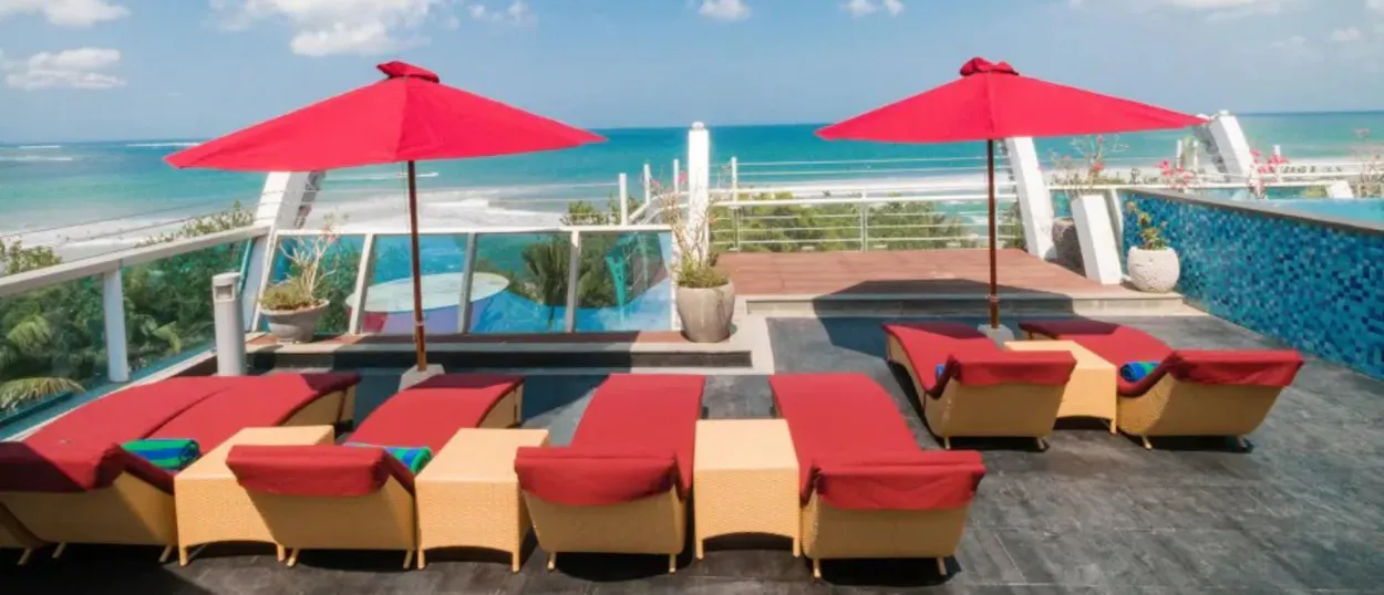 Red lounge chairs and umbrellas on a sunny rooftop terrace overlooking the ocean and Kuta Beach.