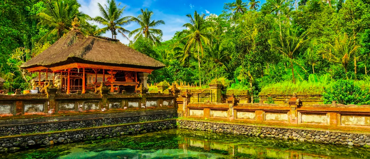 A tranquil Balinese temple with a thatched roof beside a clear pond, nestled in lush tropical foliage.