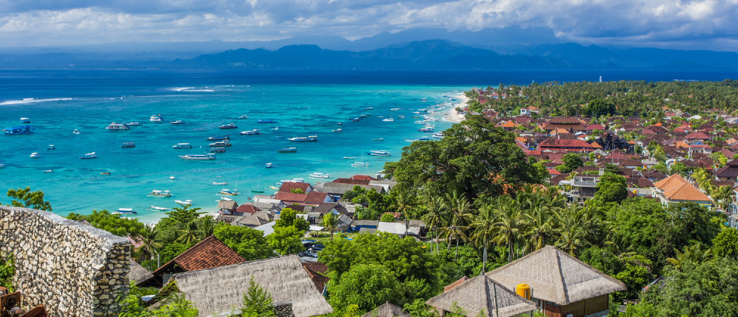 Beautiful coastal town in Bali with lush greenery, traditional roofs, and boats in turquoise ocean.