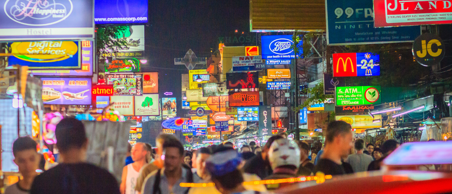 Crowds of people walk past a vibrant street lined with colorful neon signs at night in Bangkok.