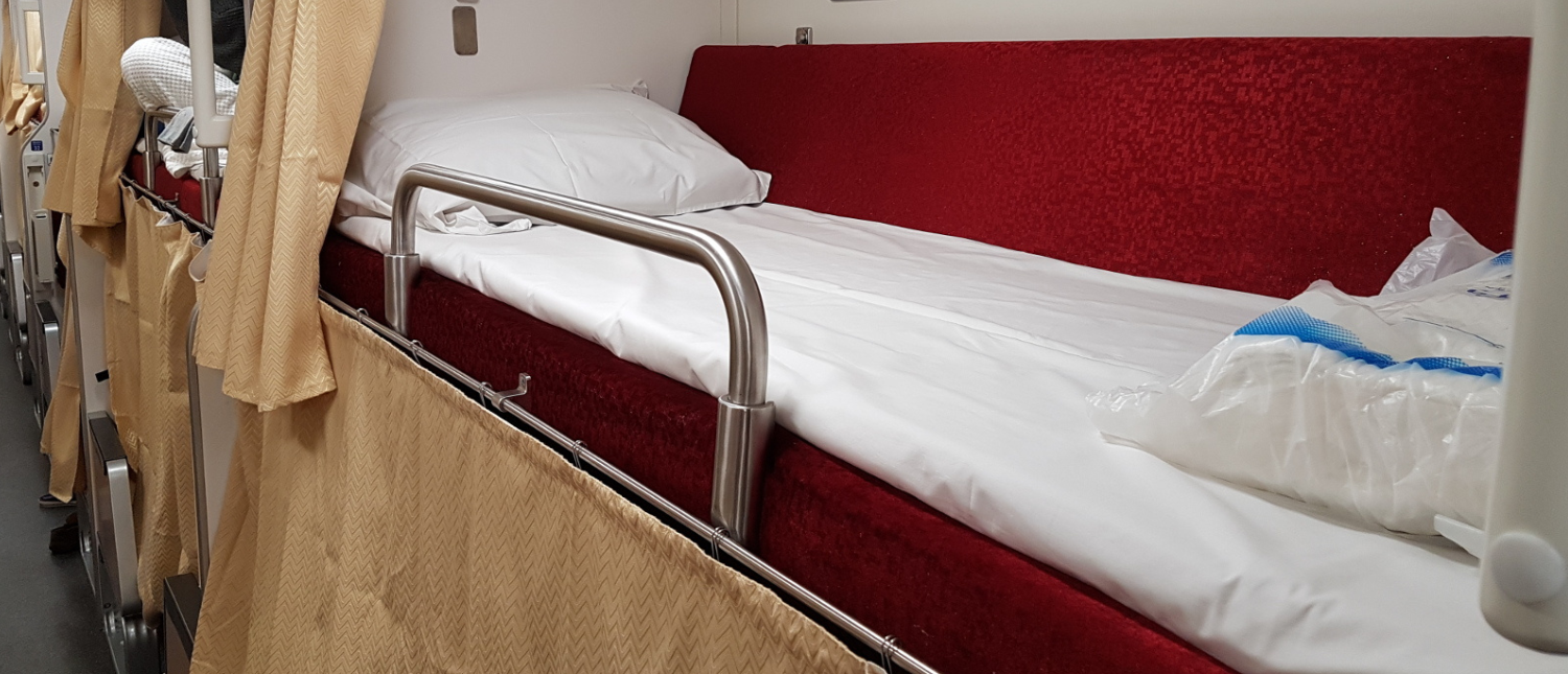Comfortable bunk bed with red mattress and white sheets inside a Thai sleeper train cabin.