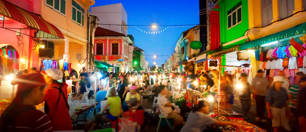 A vibrant night market in Phuket Old Town with many people browsing colorful stalls under string lights.