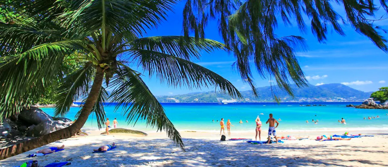 People relax on a sunny Phuket beach with white sand, turquoise water, and palm trees.