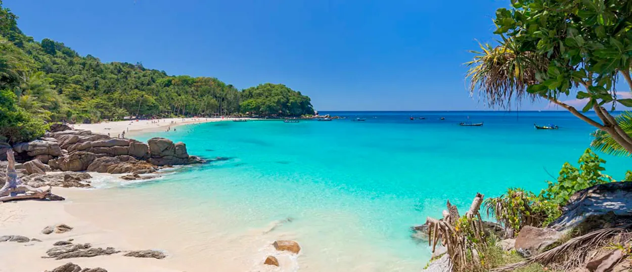 Serene white sand beach with clear turquoise water, green hills, and distant boats in Phuket.