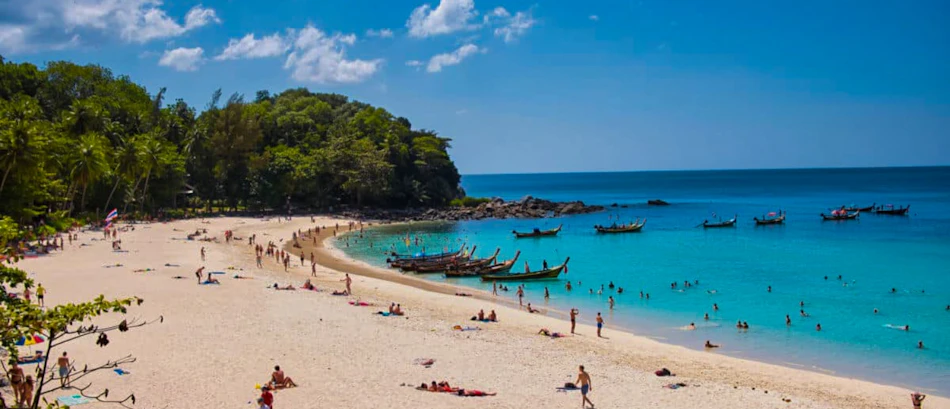 Phuket's stunning beach with sunbathers, swimmers, and traditional longtail boats floating on the clear turquoise sea.