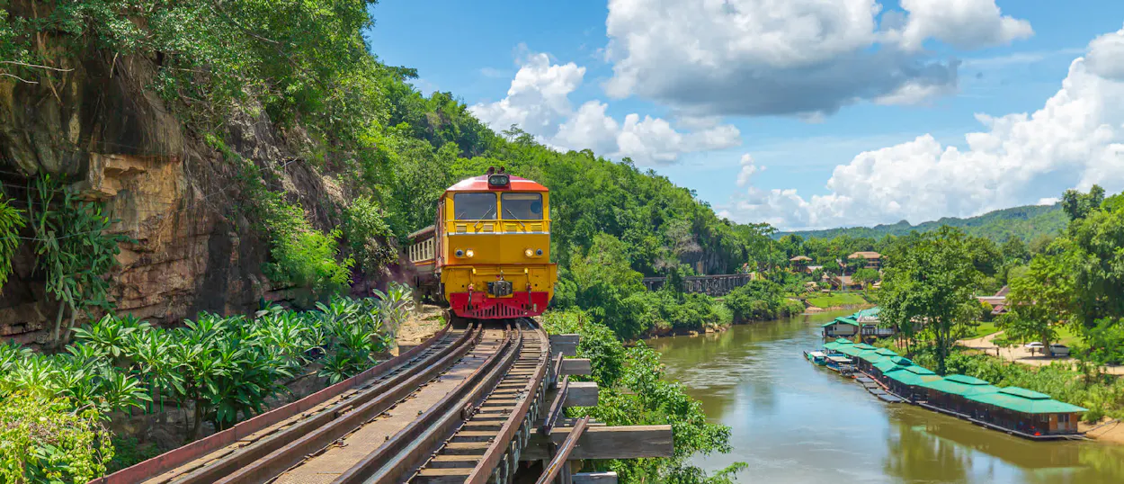 A vibrant yellow and red train travels along tracks beside a river in lush Thai countryside.