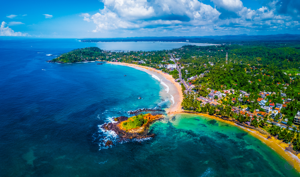 Aerial view of Weligama Bay, Sri Lanka, with a curving sandy beach and clear turquoise waters.