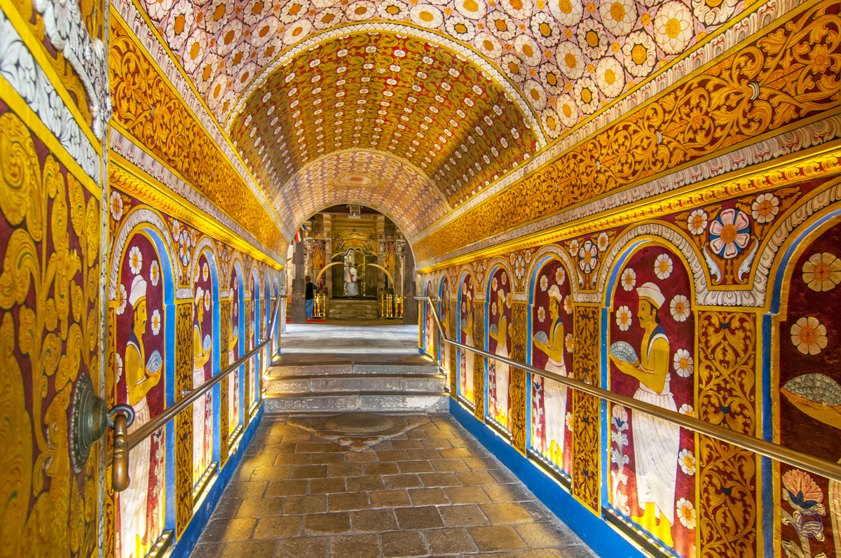A vibrant, ornate hallway with golden and maroon murals leading to ancient statues inside Dambulla Cave Temple.