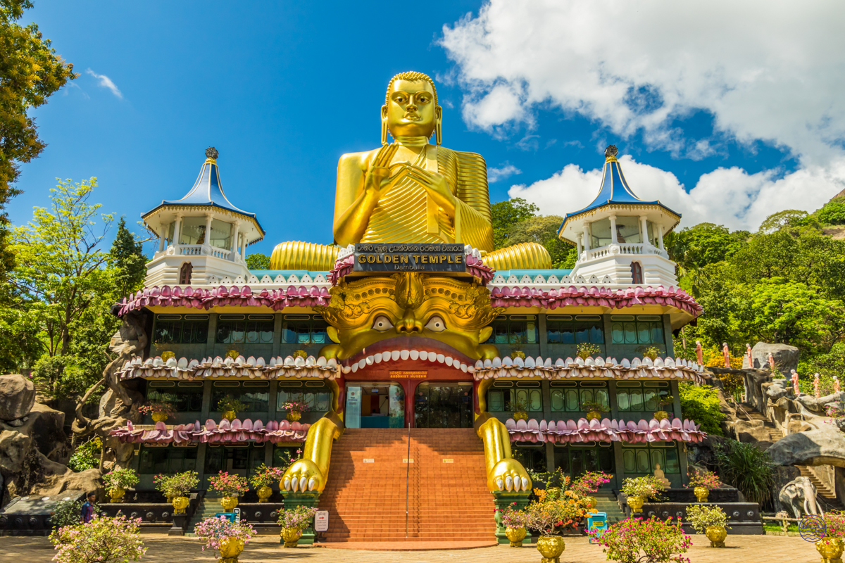 A majestic golden Buddha statue sits atop the ornate Golden Temple of Dambulla under a bright blue sky.