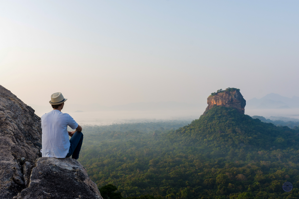 A person sits on Pidurangala Rock, admiring the majestic Sigiriya Rock fortress and lush Sri Lankan jungle.