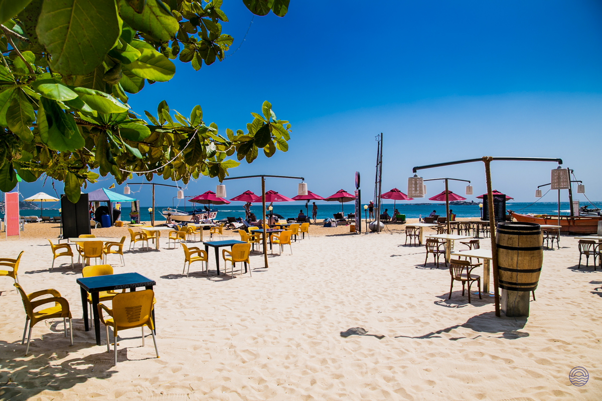 Golden sands of Weligama beach, dotted with yellow chairs and red umbrellas under a bright blue sky.