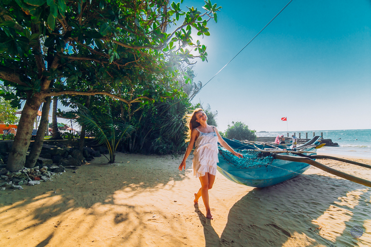 A joyful woman walks on the sunny golden beach of Weligama, past a traditional blue fishing boat.