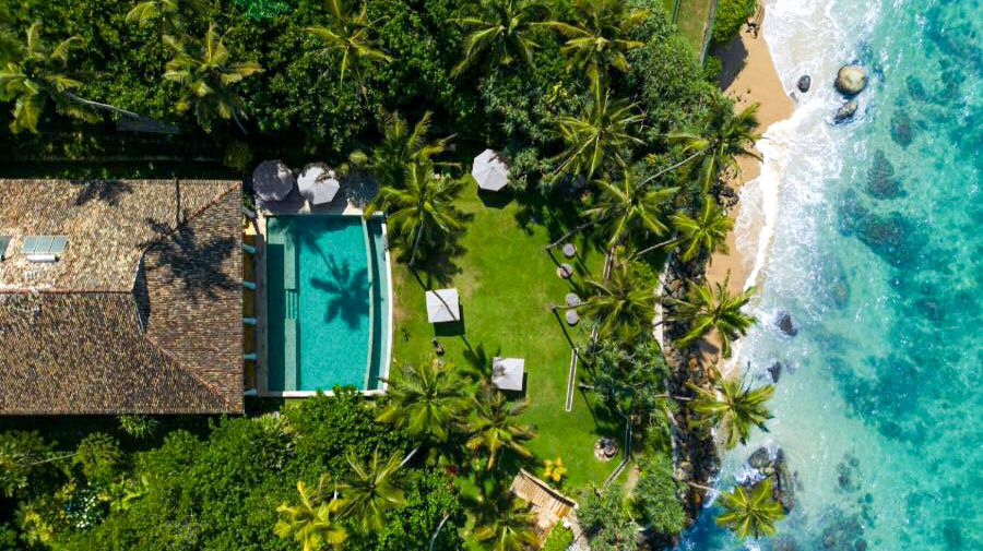 Aerial view of a luxurious villa with a pool and palm trees beside Weligama's turquoise beach.