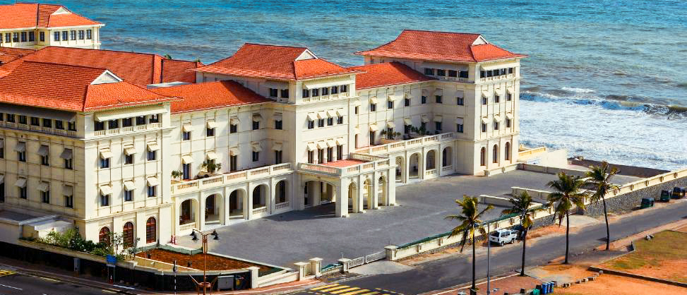 Elegant colonial-style hotel with red roofs overlooking the ocean and palm trees in Colombo, Sri Lanka.