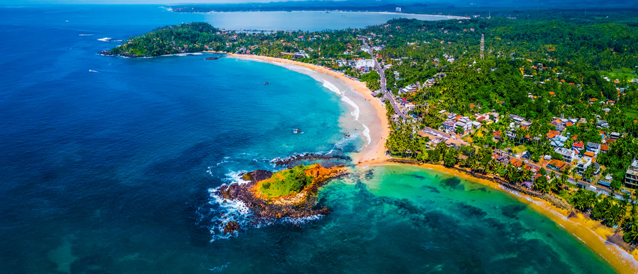 Stunning aerial view of Mirissa beach with clear blue waters, a rocky island, and lush palms.