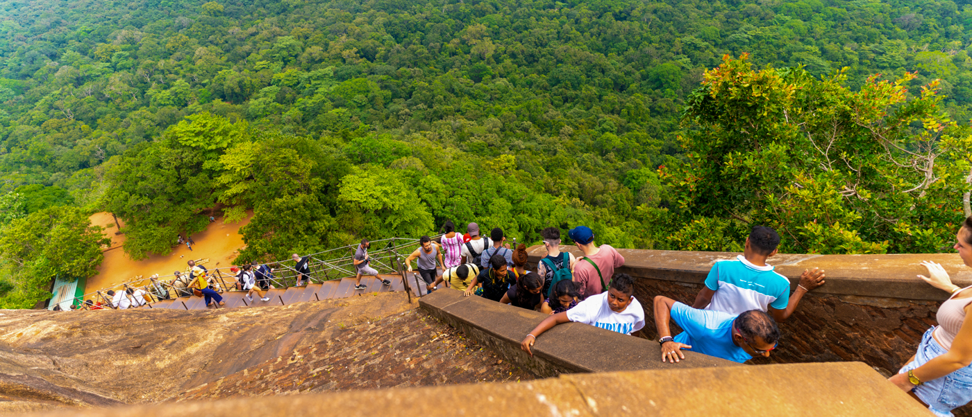 Tourists climb steep ancient steps at Sigiriya, overlooking a vast green jungle.