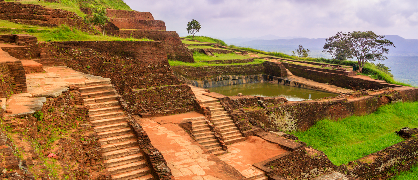 Ancient stone steps and terraced ruins of Sigiriya fortress with a pool and distant hills.