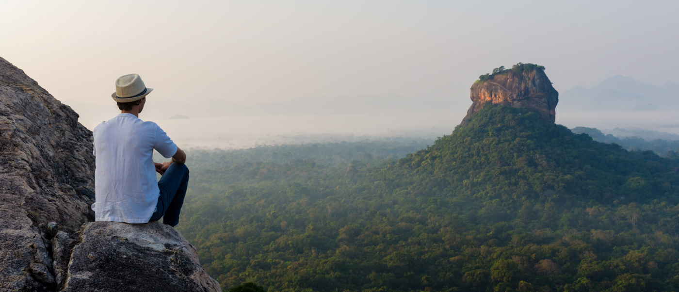 A traveler sits on Pidurangala Rock, enjoying the panoramic view of Sigiriya and the misty jungle.