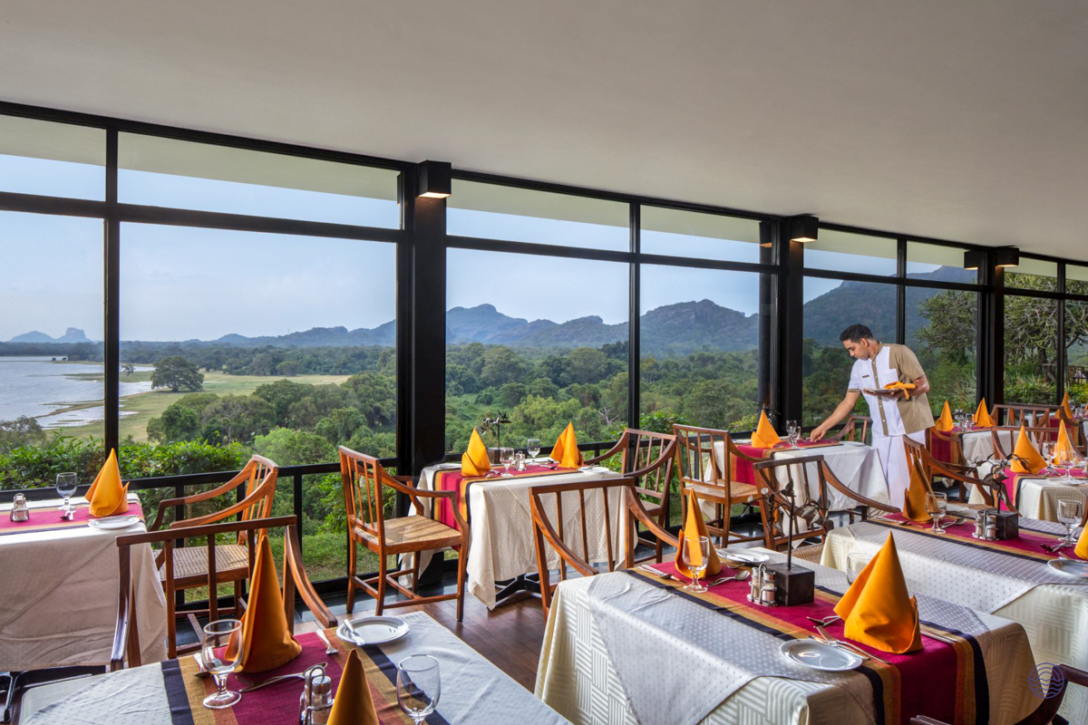 A waiter prepares tables in a scenic restaurant overlooking Kandalama Lake and lush Sri Lankan landscape.