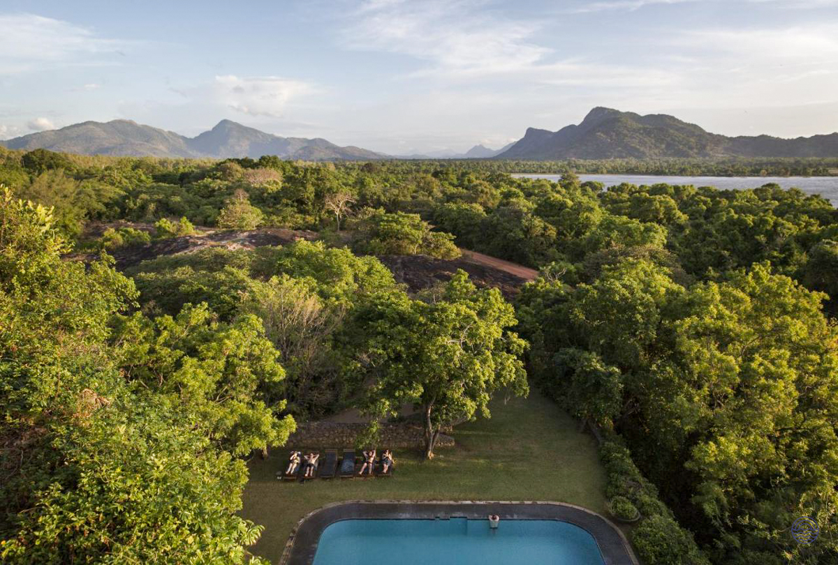 Guests relax by a resort pool surrounded by lush forest, a lake, and mountains in Sri Lanka.