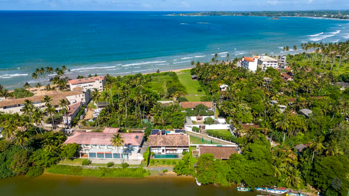 Aerial view of the tropical Weligama coastline with hotels, lush palm trees, and a river meeting the ocean.