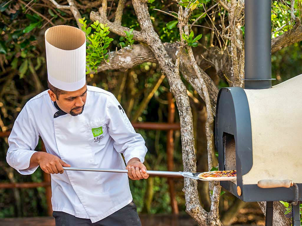 A chef expertly slides a freshly prepared pizza into an outdoor wood-fired oven at Uga Chena Huts.