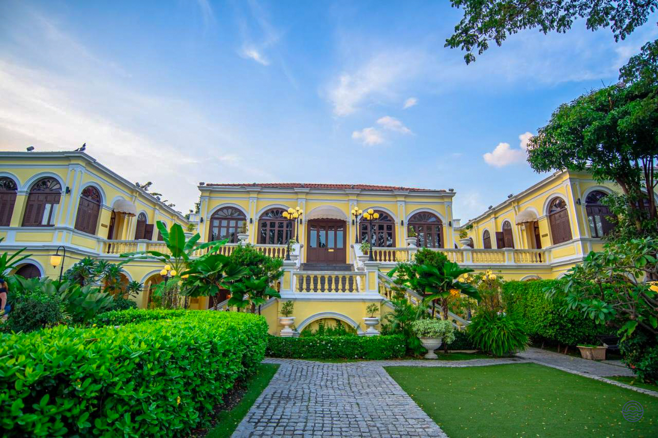 An elegant yellow historic hotel building with a grand entrance and lush green gardens in Bangkok.