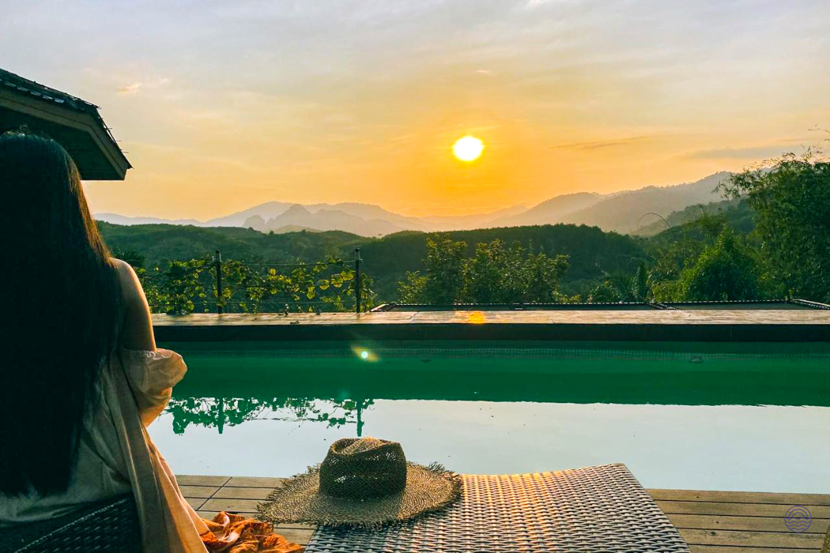 A woman relaxes by an infinity pool, enjoying a stunning sunset over the lush Khao Sok mountains.