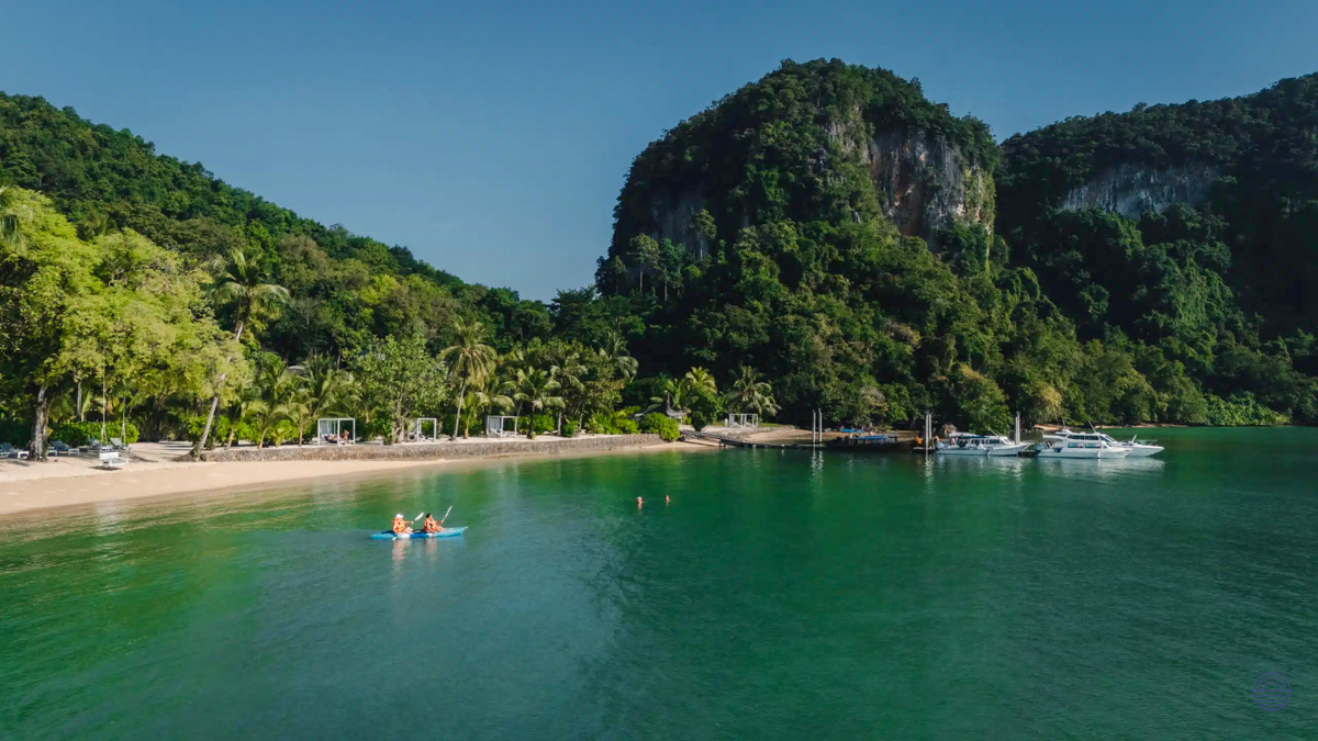 Two people kayak on turquoise water near a tropical beach and lush green mountains on Koh Yao Noi.