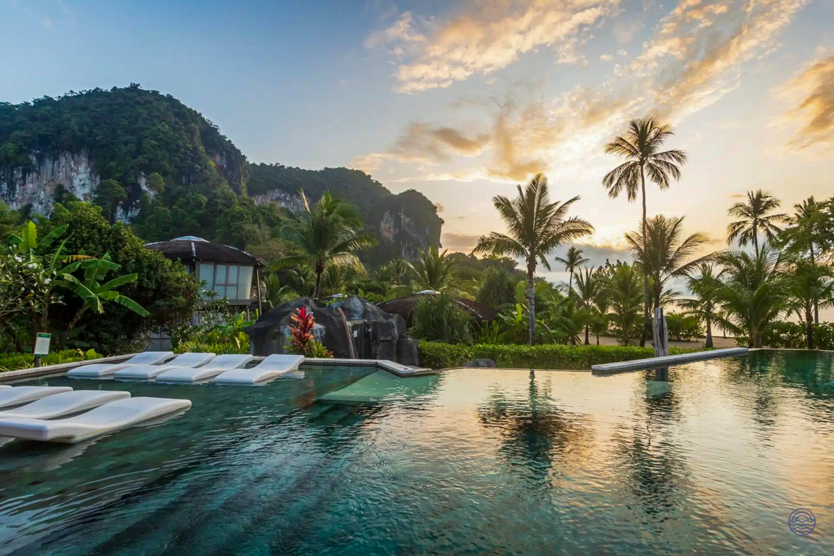 Luxurious infinity pool with white loungers, tropical mountains, and palm trees under a sunset sky in Thailand.