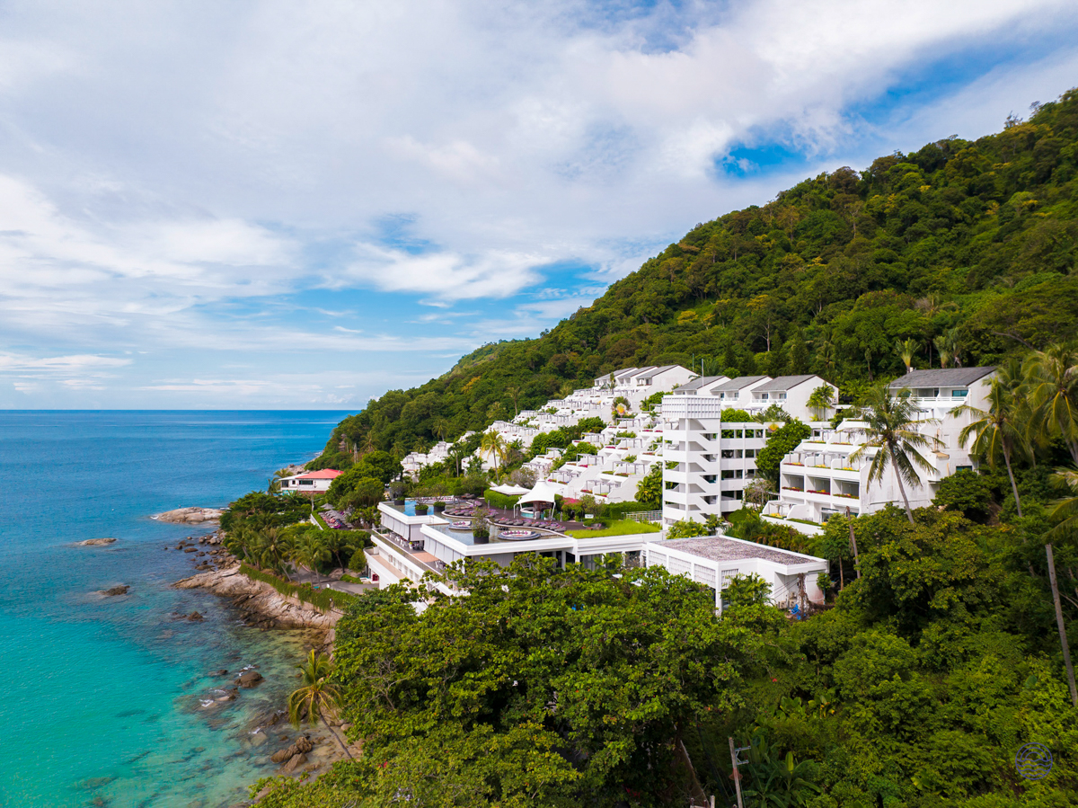 A luxurious white resort nestled into a lush green hillside overlooking the turquoise ocean in Phuket.