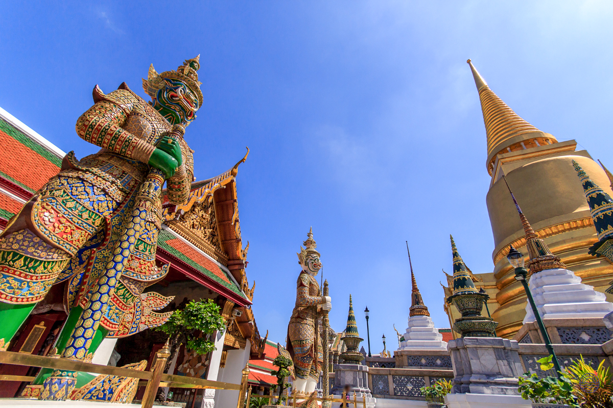 Ornate guardian statues and golden stupas stand majestically under a clear blue sky at a temple in Bangkok.