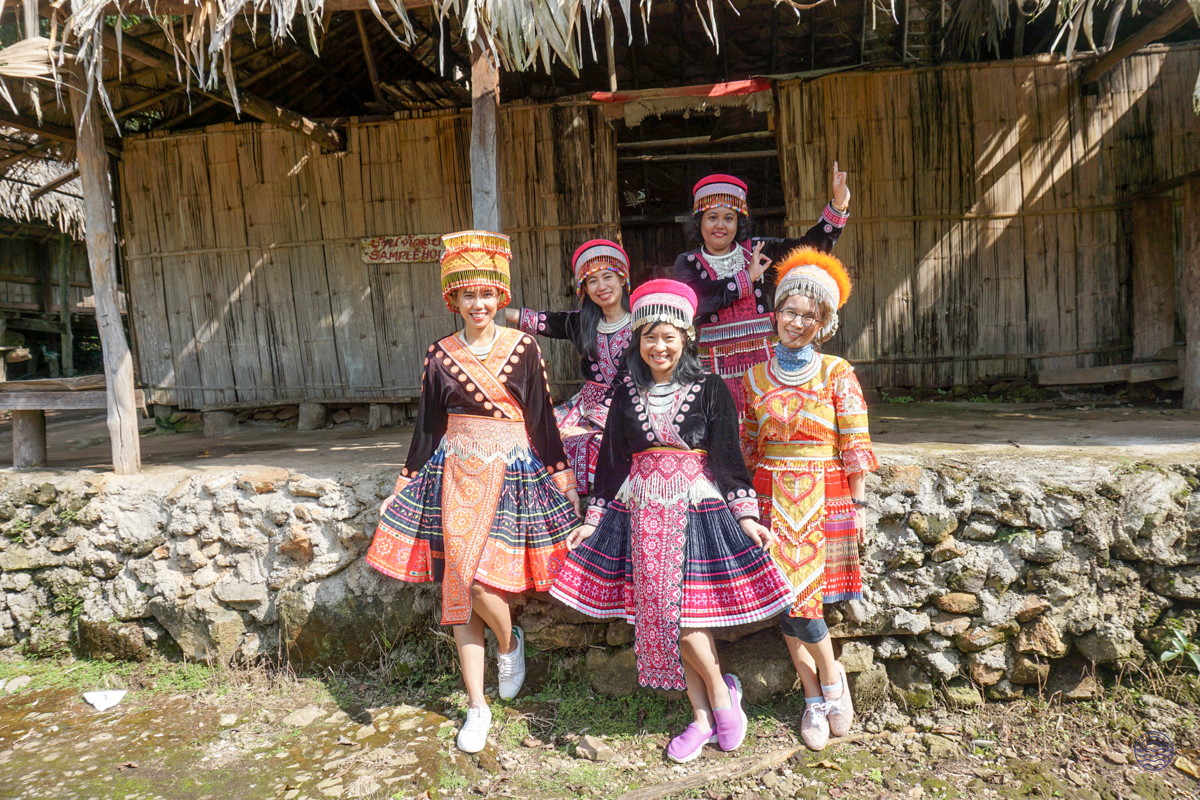Five smiling Hmong women in vibrant traditional dresses pose in their charming village near Chiang Mai.