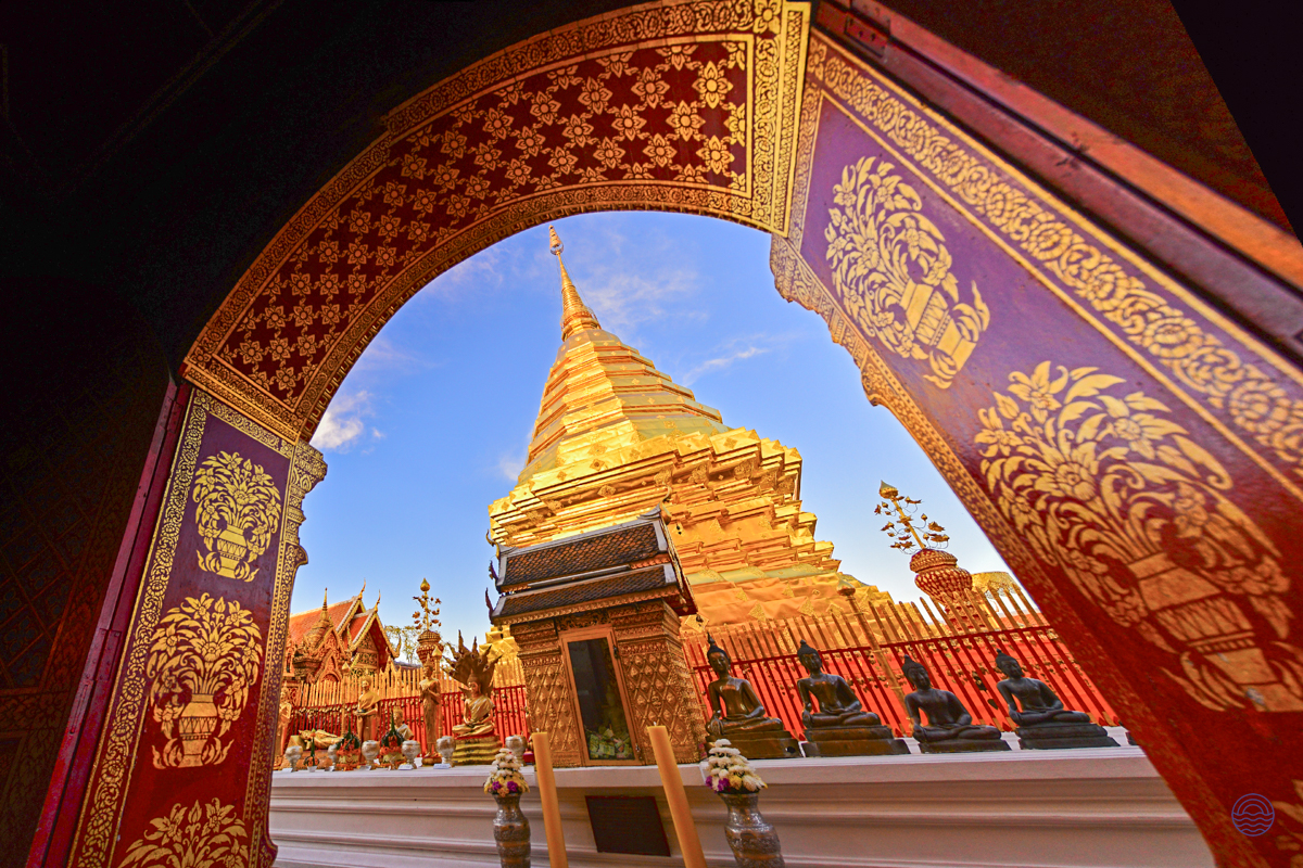 Ornate archway frames the majestic golden pagoda of Doi Suthep temple under a clear blue sky.