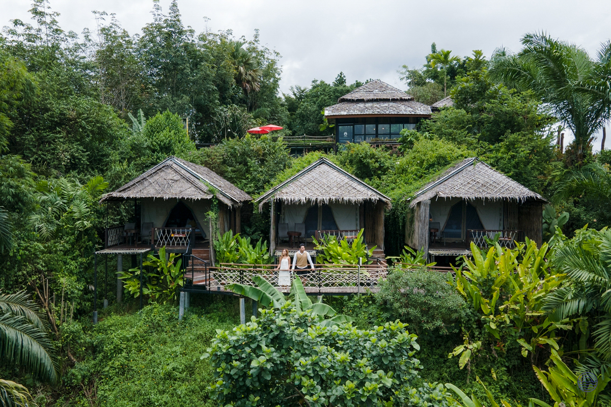 Three charming thatched bungalows in the lush green jungle of Khao Sok, with a person relaxing on a deck.