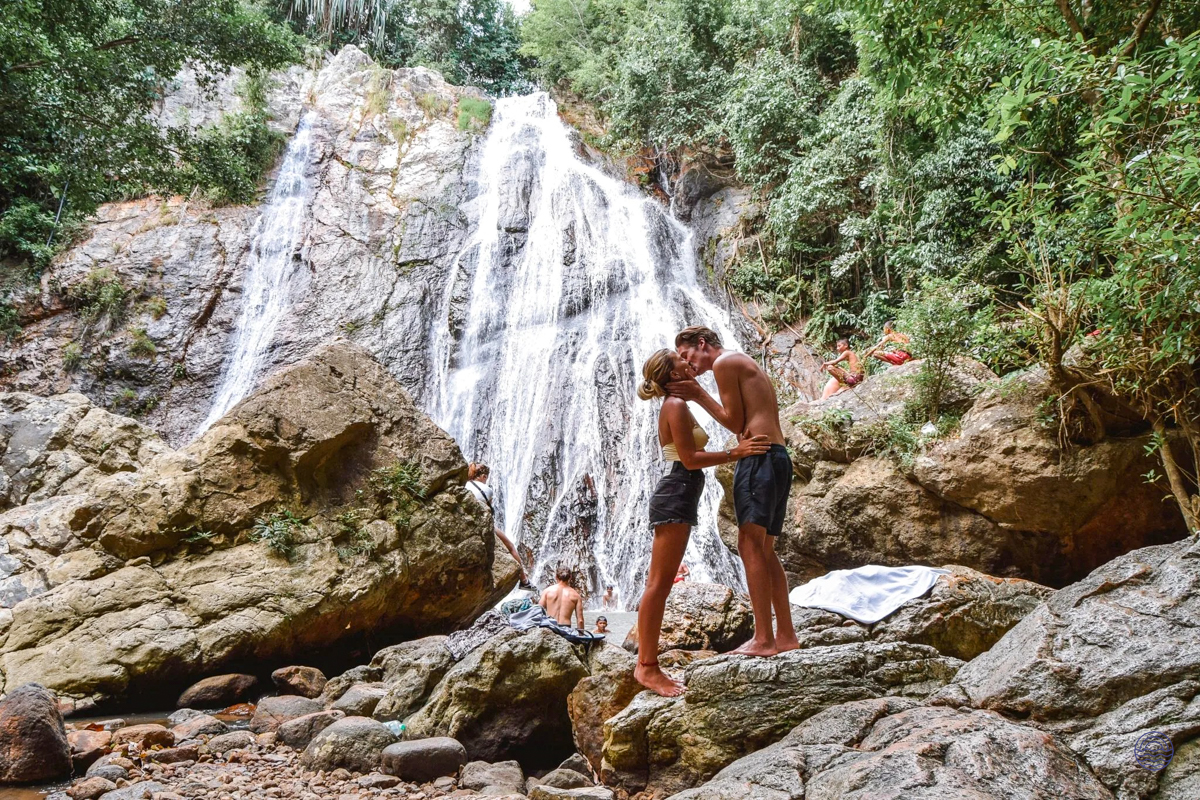 A loving couple shares a kiss on rocks before a majestic waterfall on Koh Samui.